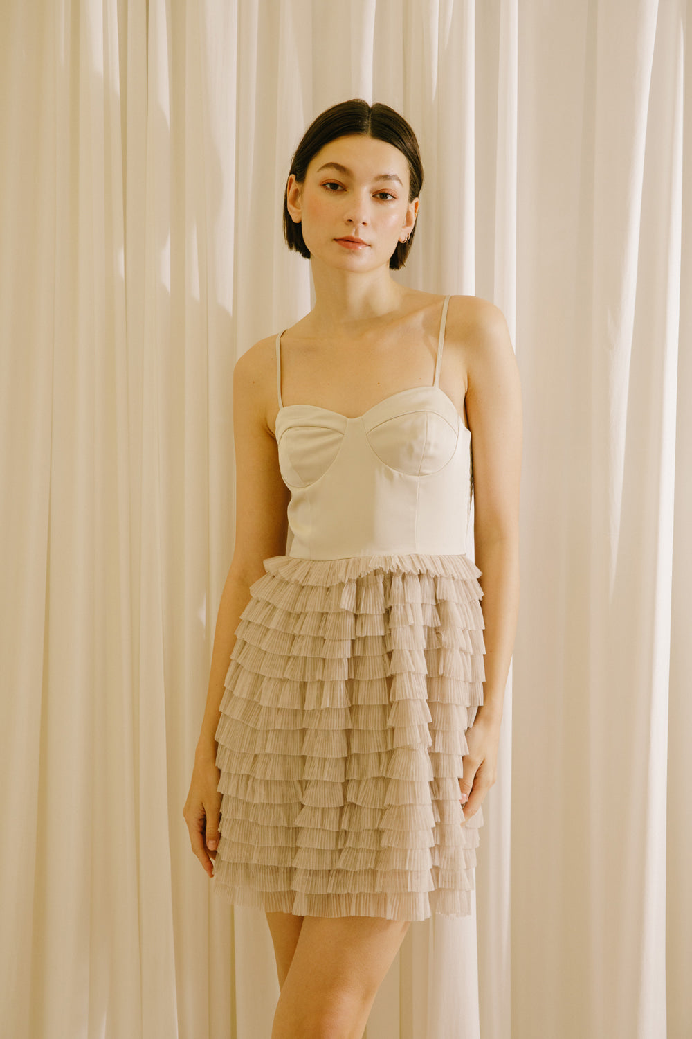 Woman wearing a beige dress with a ruffled skirt against a white curtain background