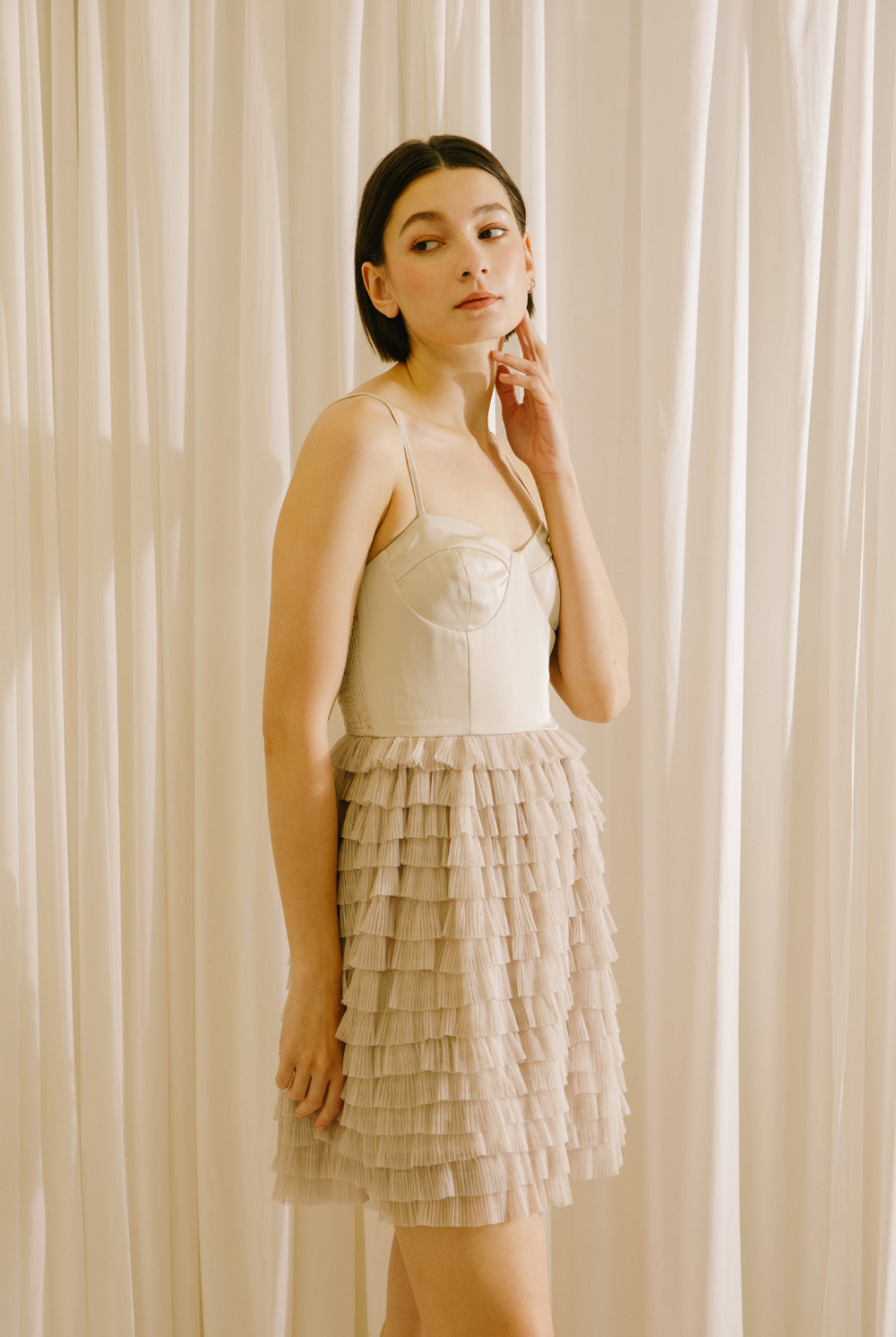 Side of a woman wearing a beige dress with a ruffled skirt against a white curtain background