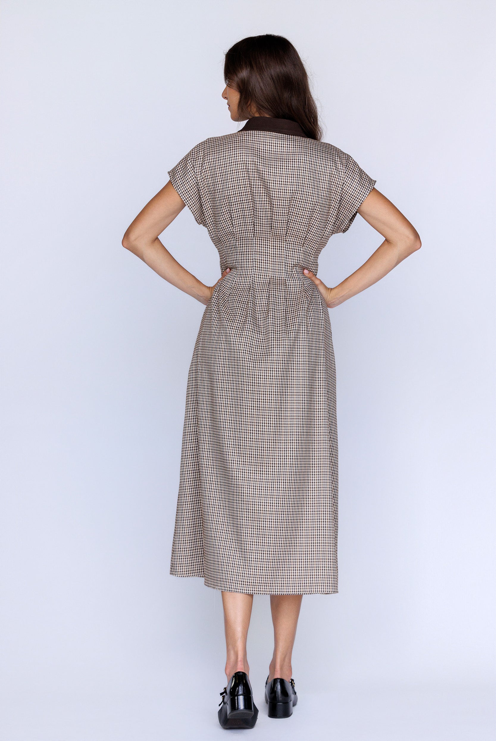 Back view of a woman wearing a brown plaid dress with her hands on her hips against a white background