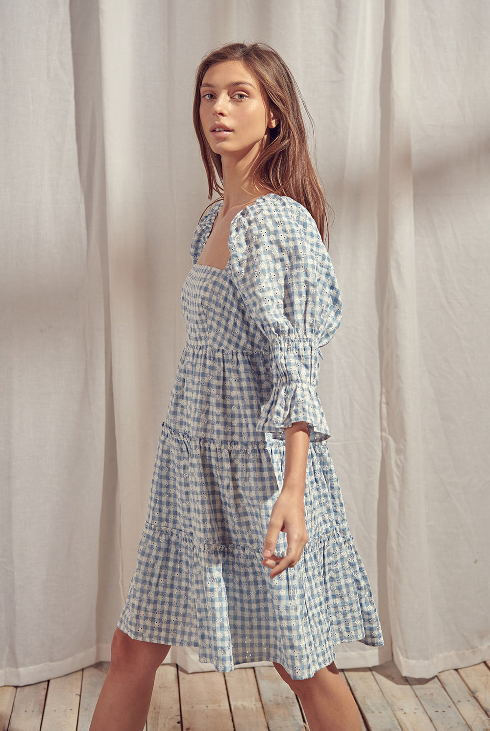Side view of a woman wearing a blue and white checkered dress with a backless design, standing on a wooden floor with a light curtain background.