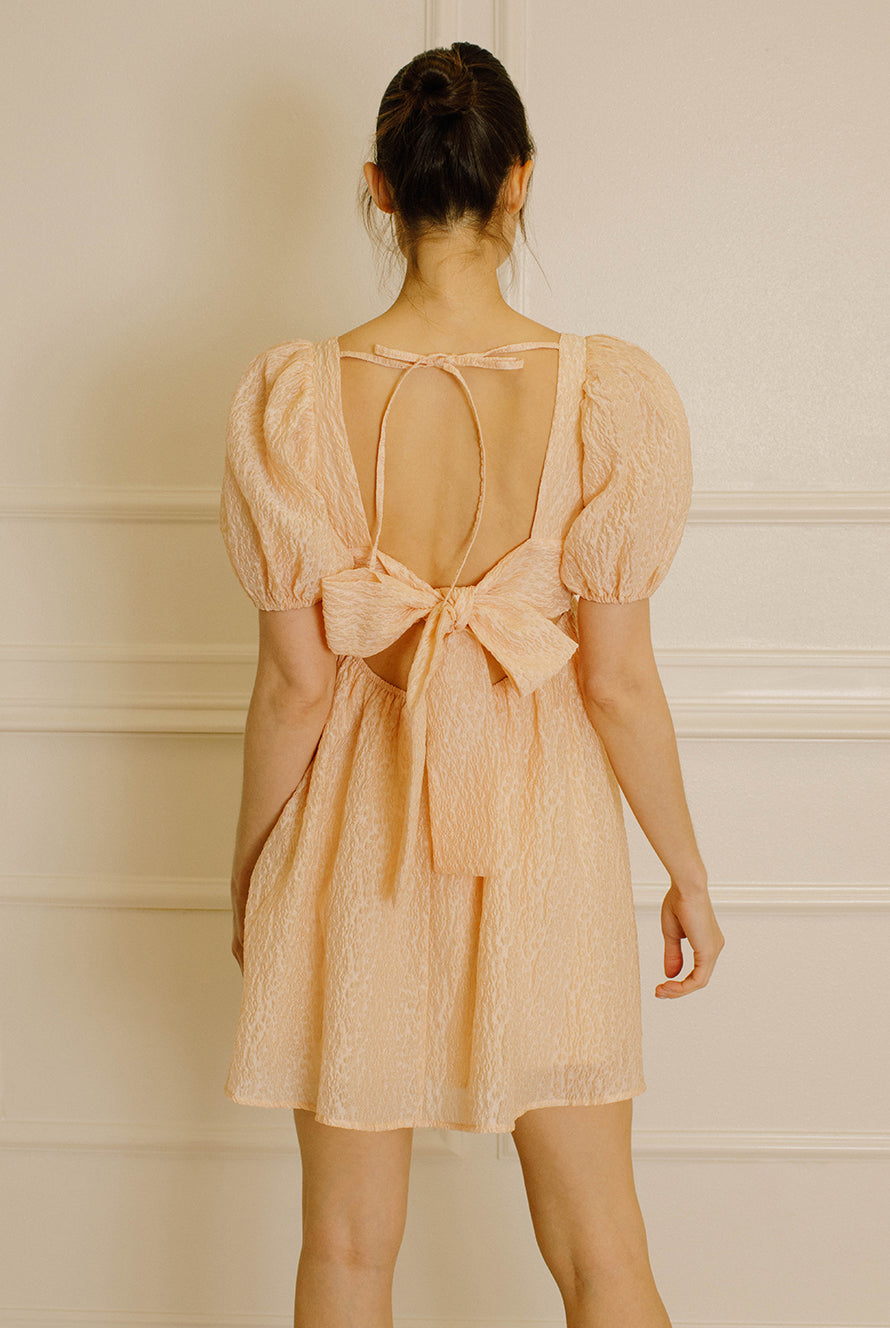 Woman wearing a peach-colored dress with an open back with two self-tie bows and beige high-heels standing against a white paneled wall
