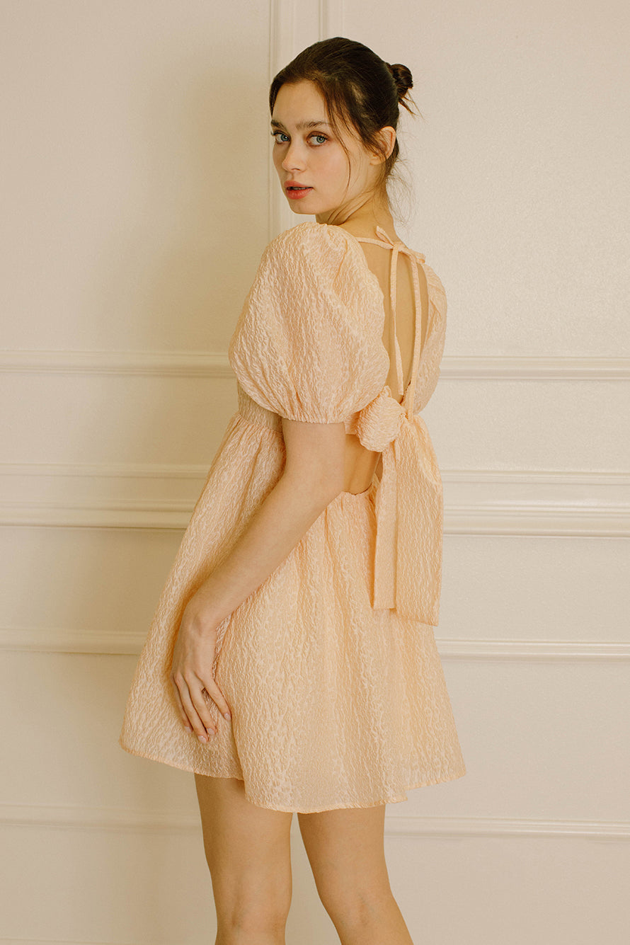 Side view of a woman wearing a peach-colored dress with an open back with two self-tie bows and beige high-heels standing against a white paneled wall