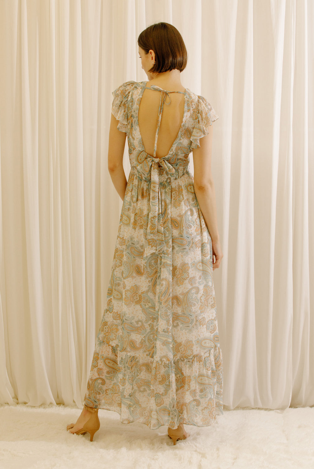 Back view of a woman wearing a beige floral dress with an open back and bow-back tie standing against a plain background