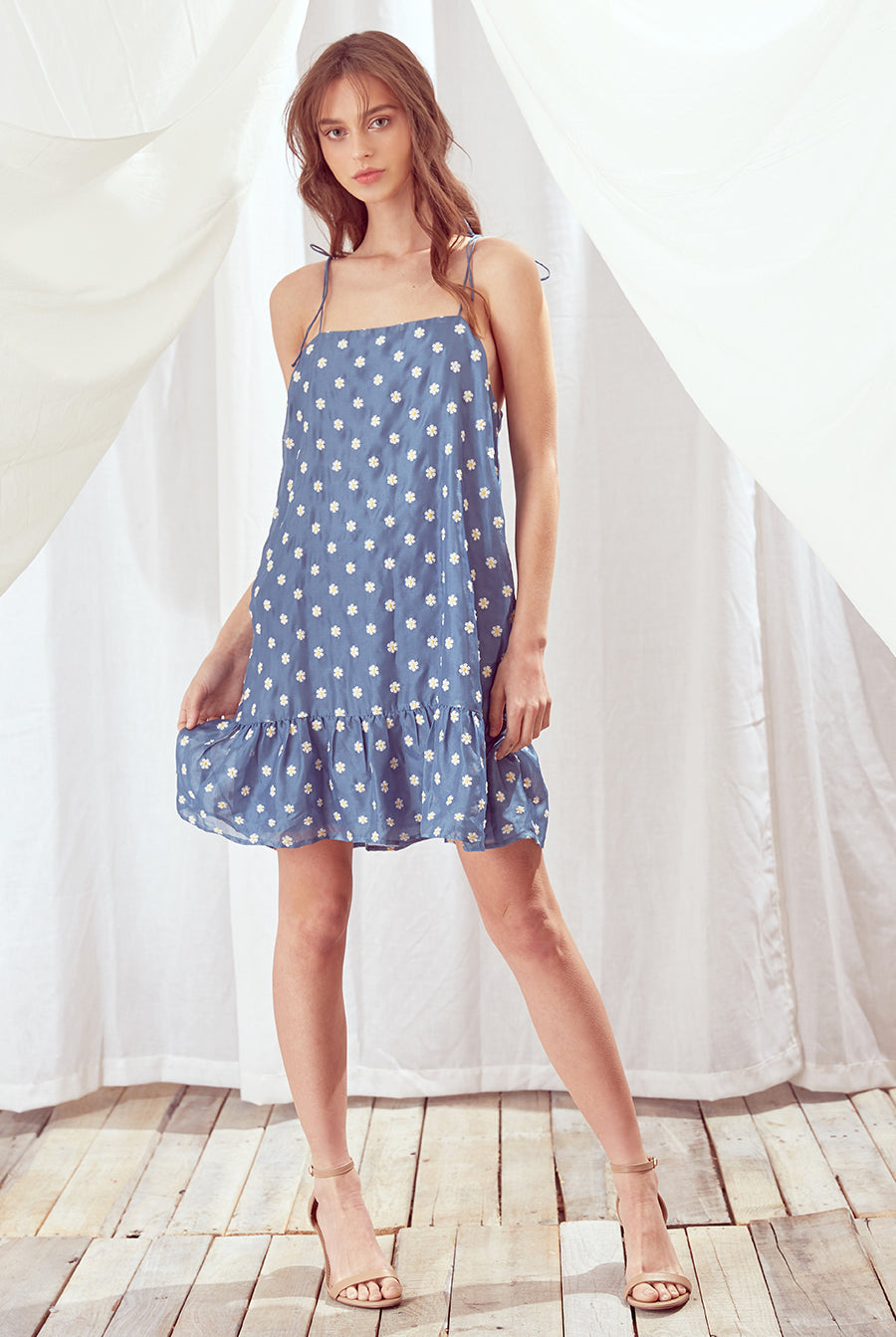 A woman standing in a room with a white and blue embroidered mini dress with daisy print, featuring spaghetti straps and a trapeze silhouette.