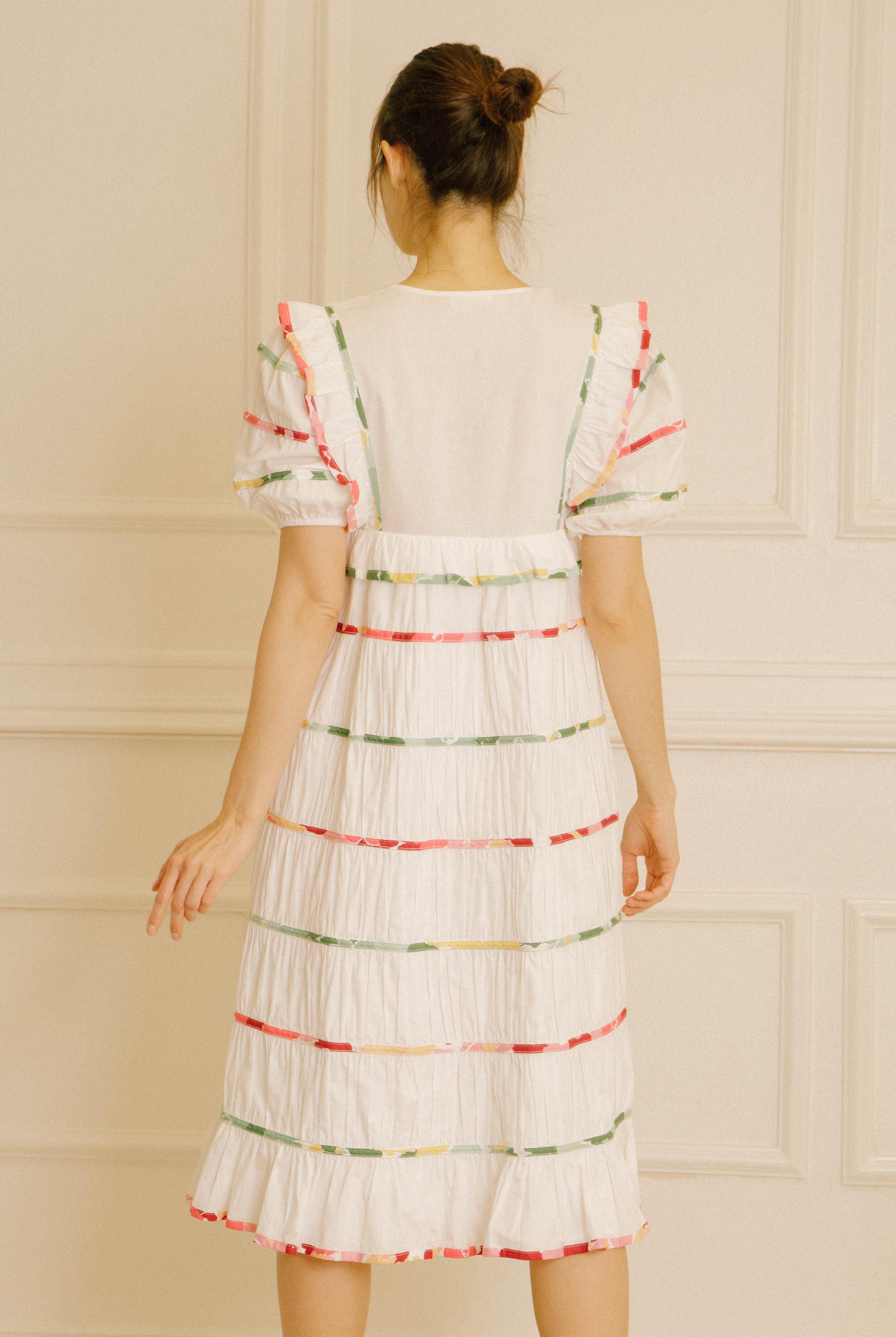 Back view of a woman wearing a white dress with colorful stripes against a beige wall.