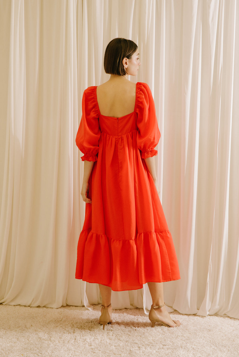 Back view of a woman wearing a red midi flowy dress standing against a white curtain backdrop