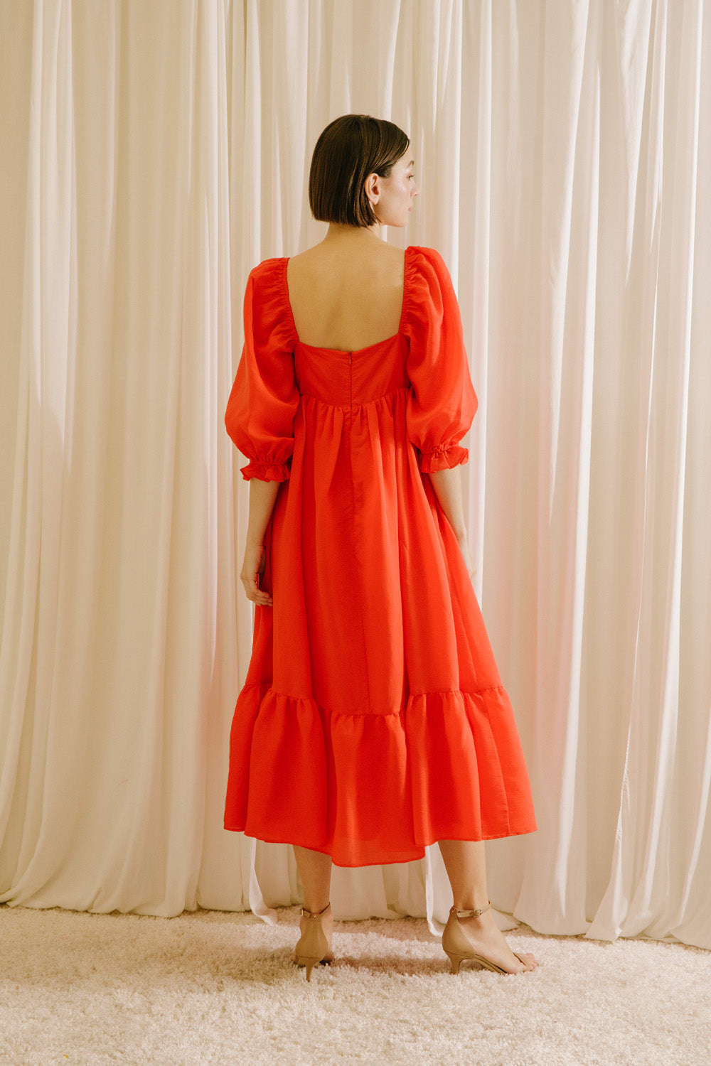 Back view of a woman wearing a red midi flowy dress standing against a white curtain backdrop