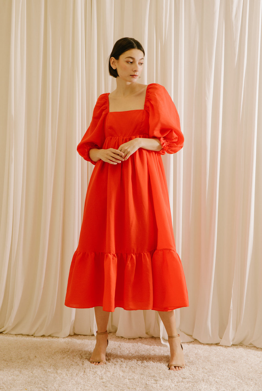 Woman wearing a red midi flowy dress standing against a white curtain backdrop
