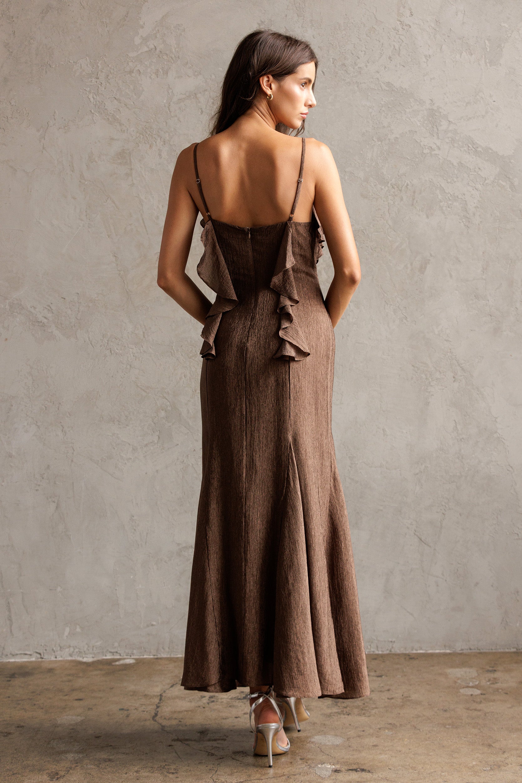 Woman wearing a brown dress with ruffled details, standing against a plain wall.