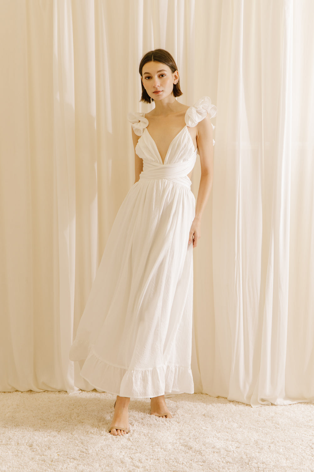 A woman wearing a white maxi dress with a plunging neckline, ruffled straps, and an empire waist. The dress features an open back with a criss-cross tie and a back zipper.