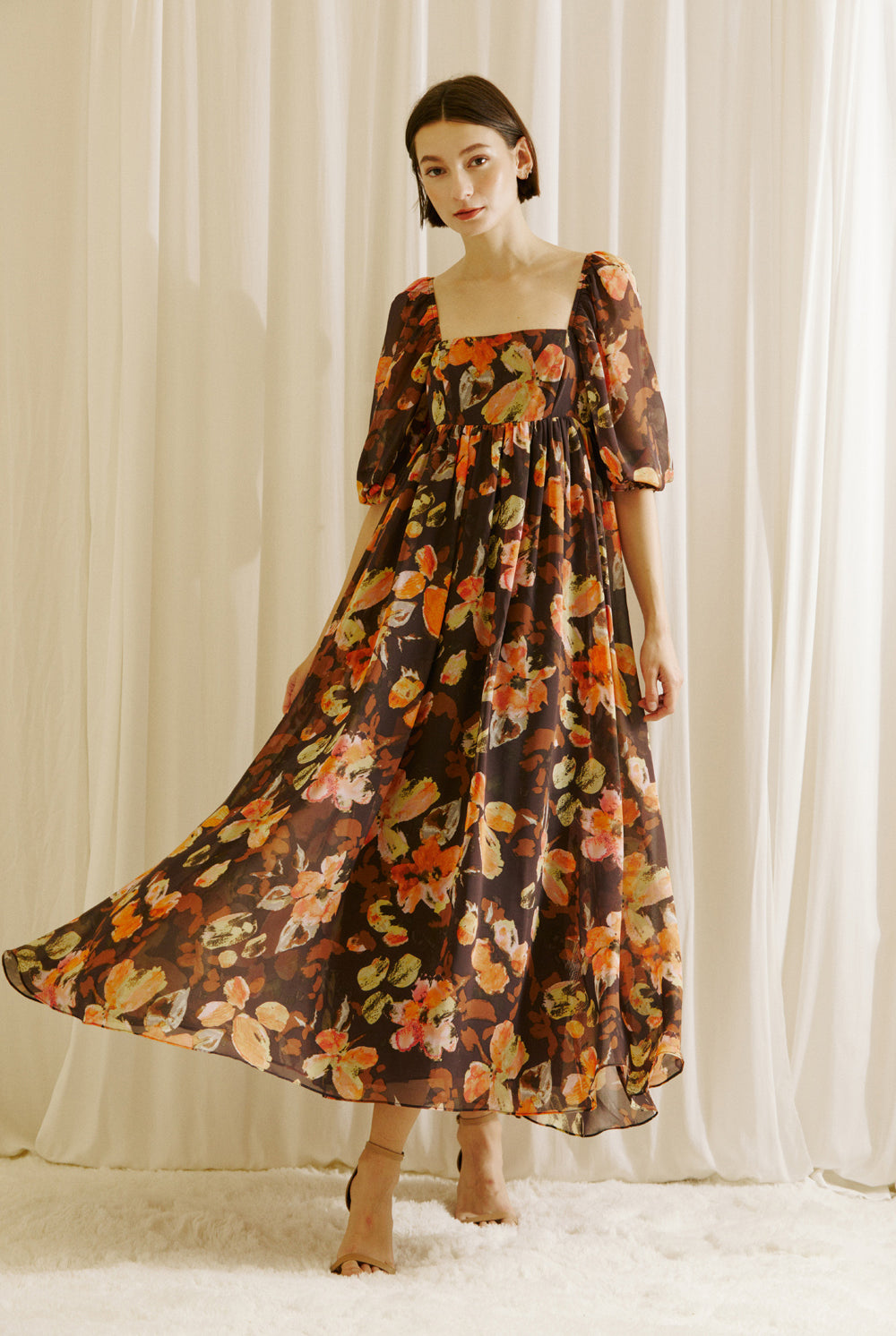 Woman wearing a brown floral dress and beige shoes standing against a white curtain backdrop