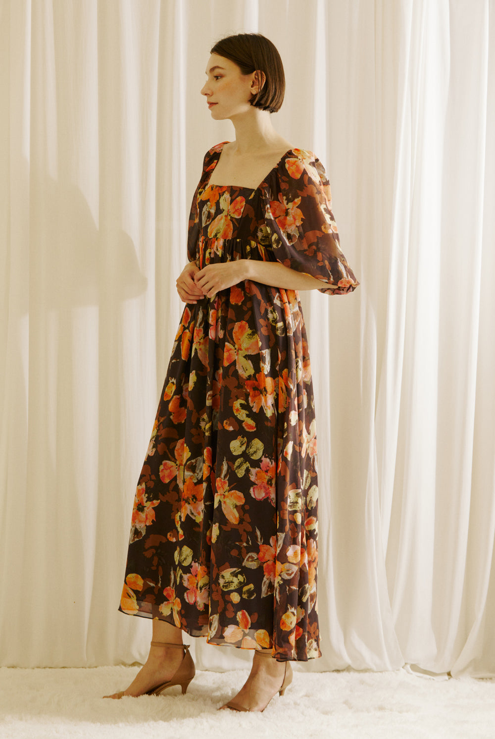 Side view of a woman wearing a brown floral dress standing against a white curtain backdrop