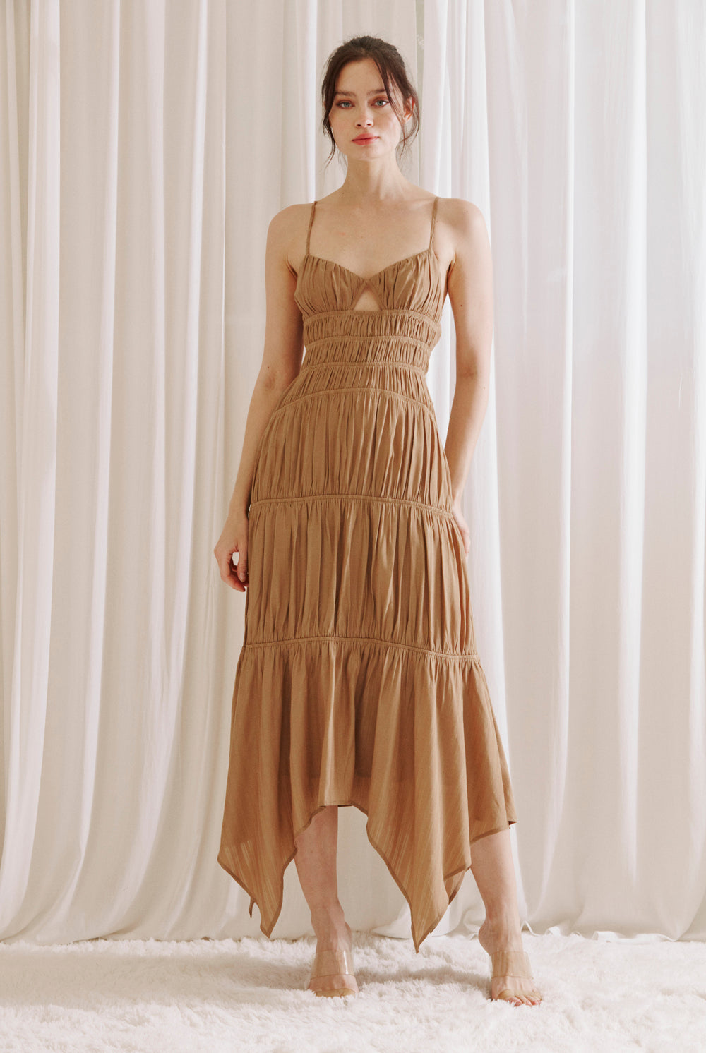 Woman wearing a brown dress with asymmetrical Hem standing against a white curtain background