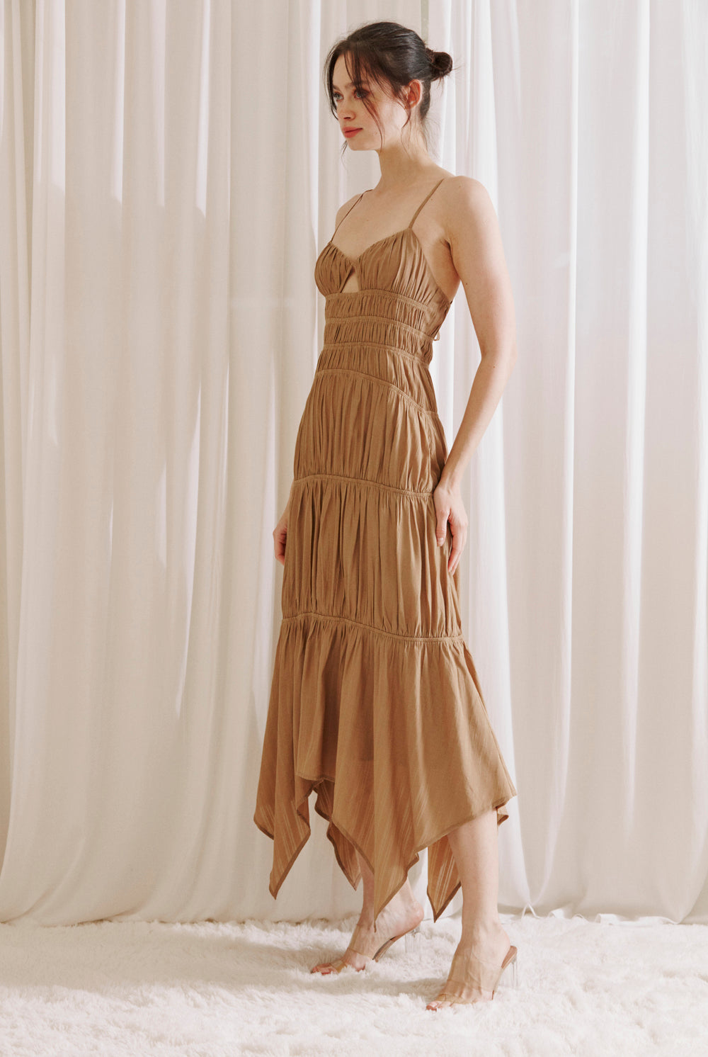 Side view of a woman wearing a brown dress with asymmetrical hem standing against a white curtain background
