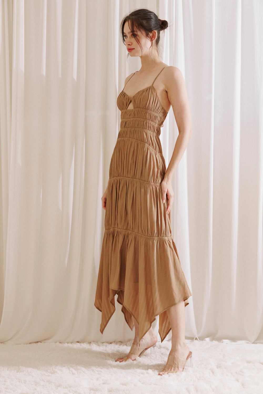 Side view of a woman wearing a brown dress with asymmetrical hem standing against a white curtain background