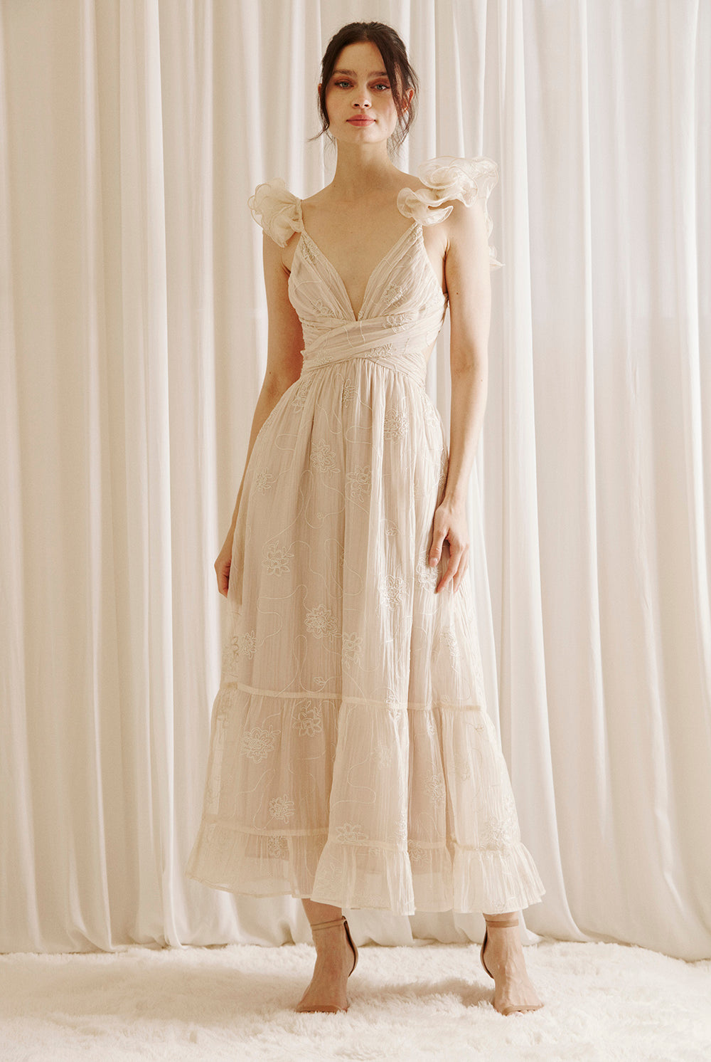 A woman wearing a beige maxi dress with embroidered flowers, featuring a plunging neckline and ruffled straps.