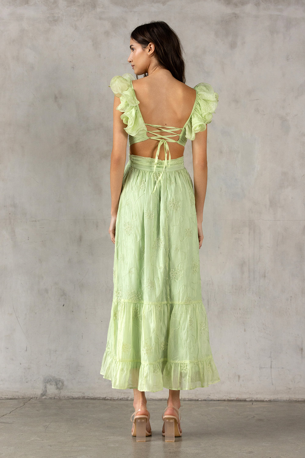 Back view of a woman wearing a lime green dress with ruffled sleeves and open back against a gray concrete wall.