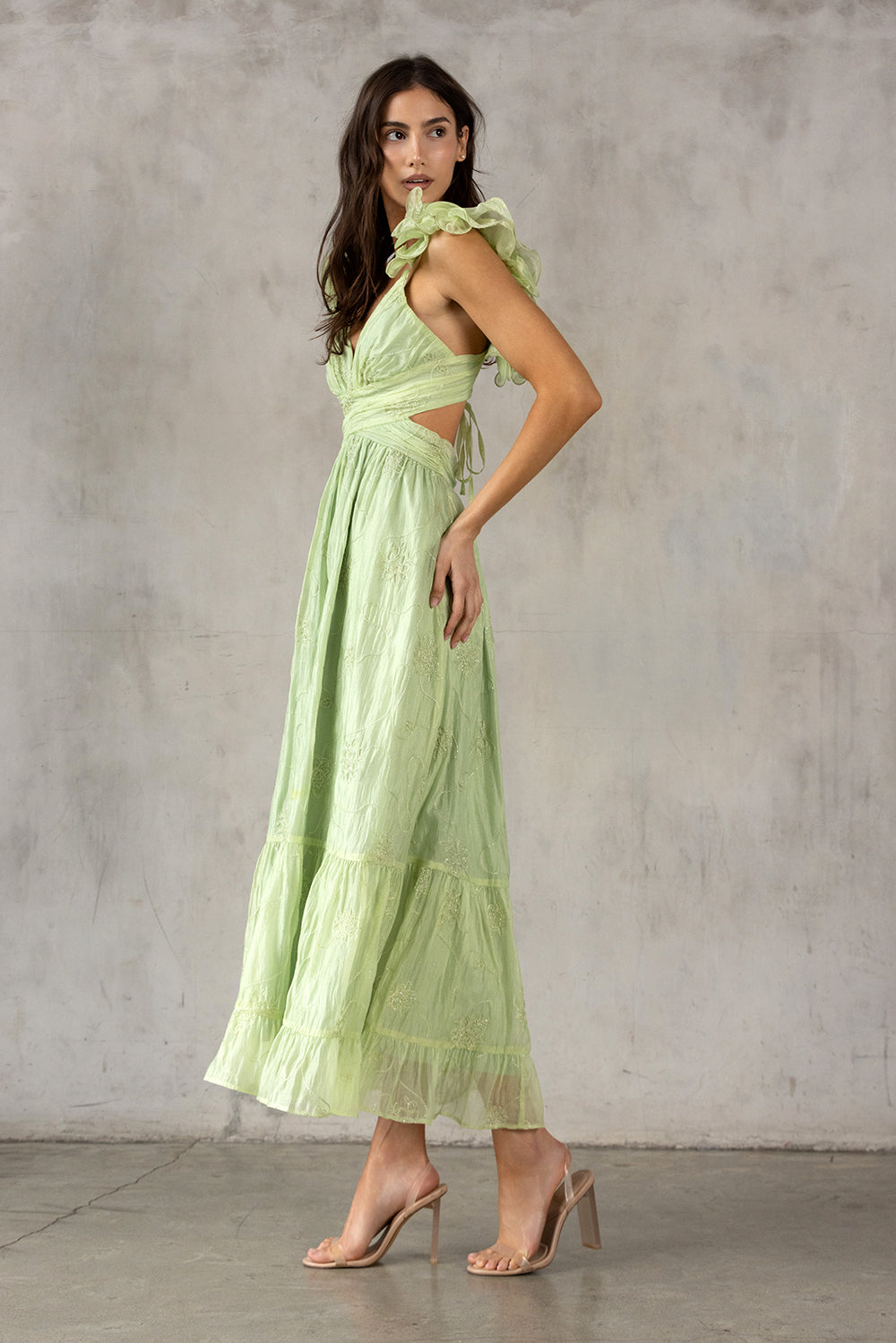 Side view of a model wearing a lime green dress with ruffled details against a gray concrete wall.