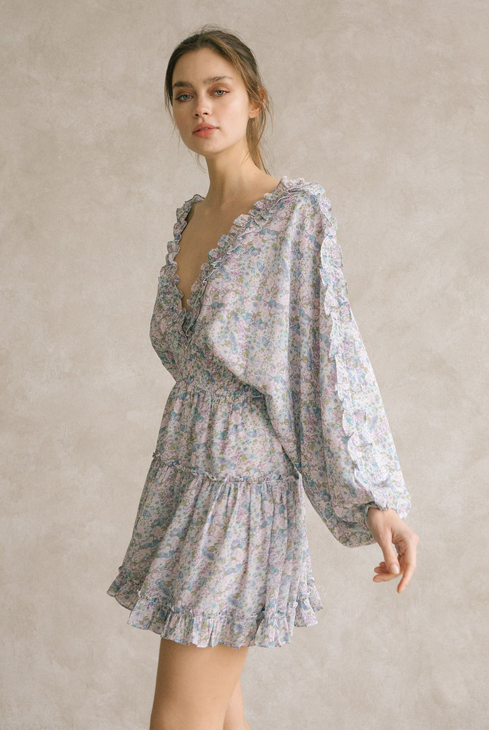 Side view of a woman wearing a blue dress with floral pattern and v-neckline with ruffle trimming against a neutral background