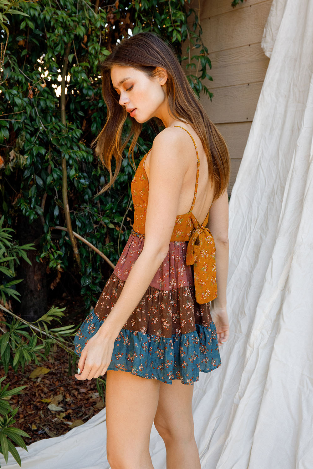 Woman wearing a floral tiered dress standing outdoors near greenery and a wooden structure.