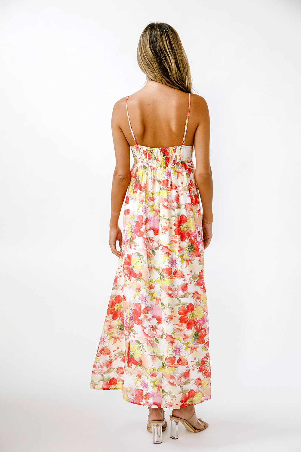 A woman viewed from behind wearing a sleeveless maxi dress with thin straps and a colorful floral print on a light cream background. She is standing against a plain white studio background.