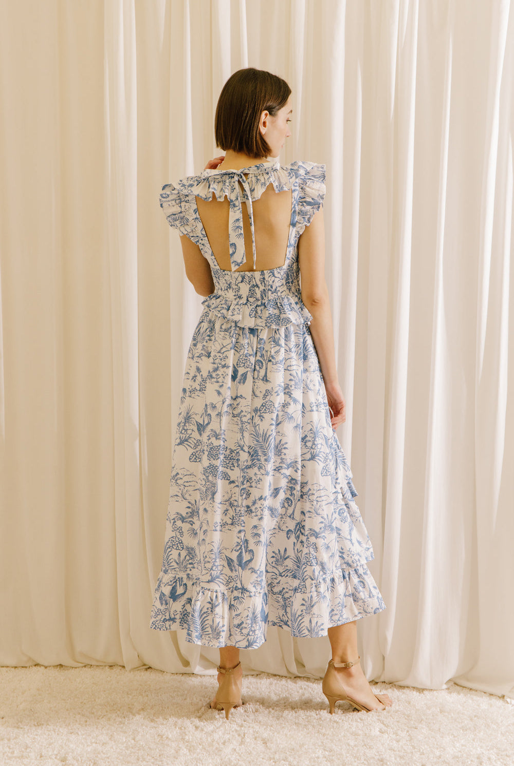 Back view of a woman wearing a white and blue tropical print midi dress standing against a beige curtain.