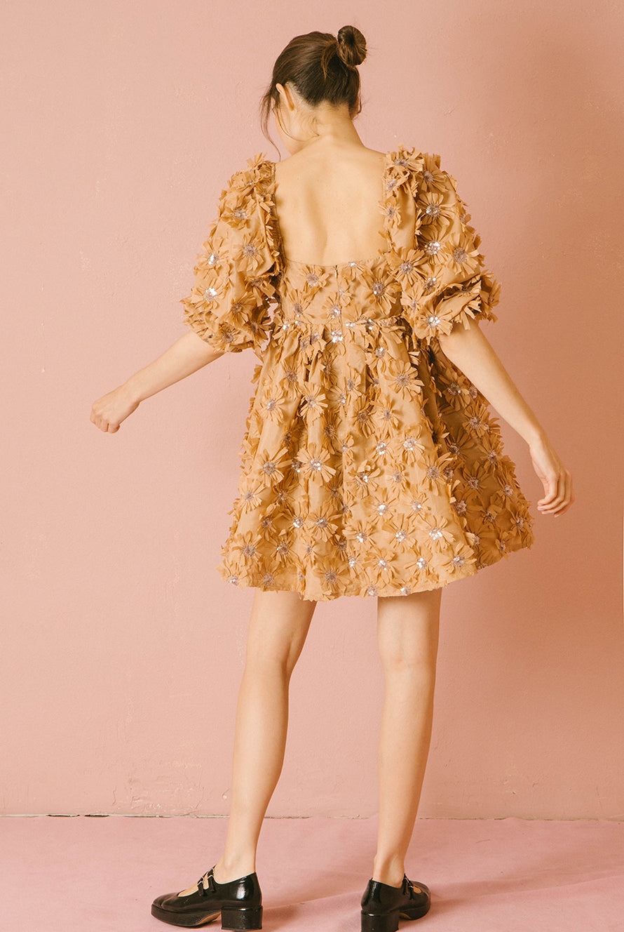 Back view of a woman wearing a caramel dress with 3D floral detail throughout against a pink background