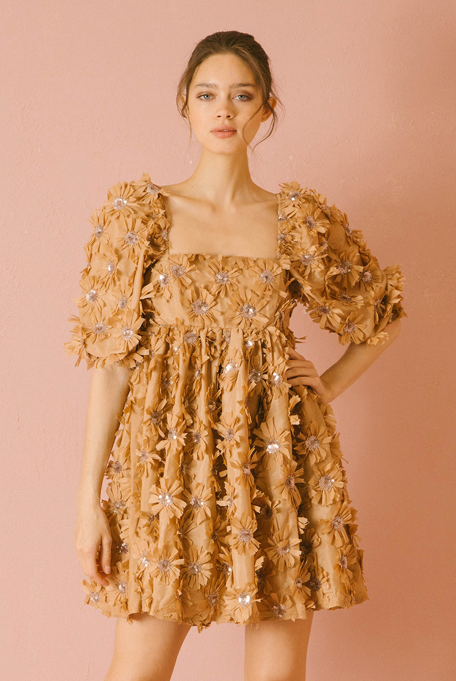Woman wearing a caramel dress with 3D floral detail throughout against a pink background