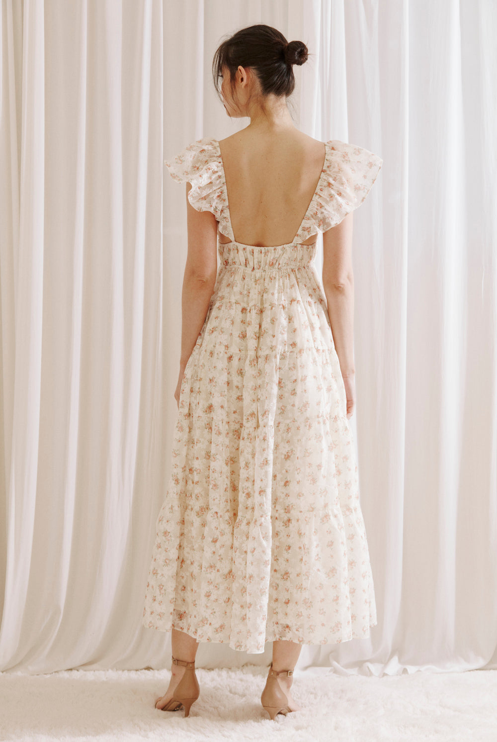 Back view of a woman wearing a floral maxi dress against a white curtain background