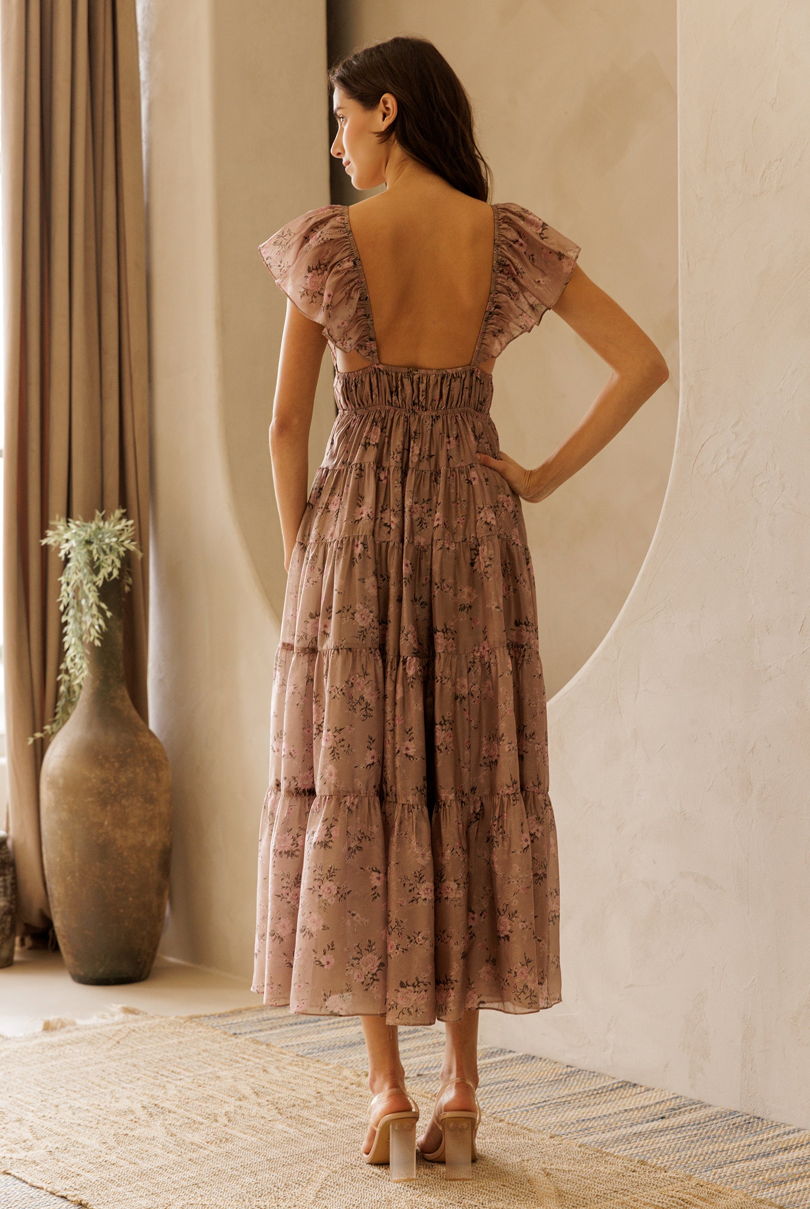 Back view of a model wearing a mauve midi dress with a floral pattern, flowy A-line silhouette, open back and tiered skirt against a beige wall.