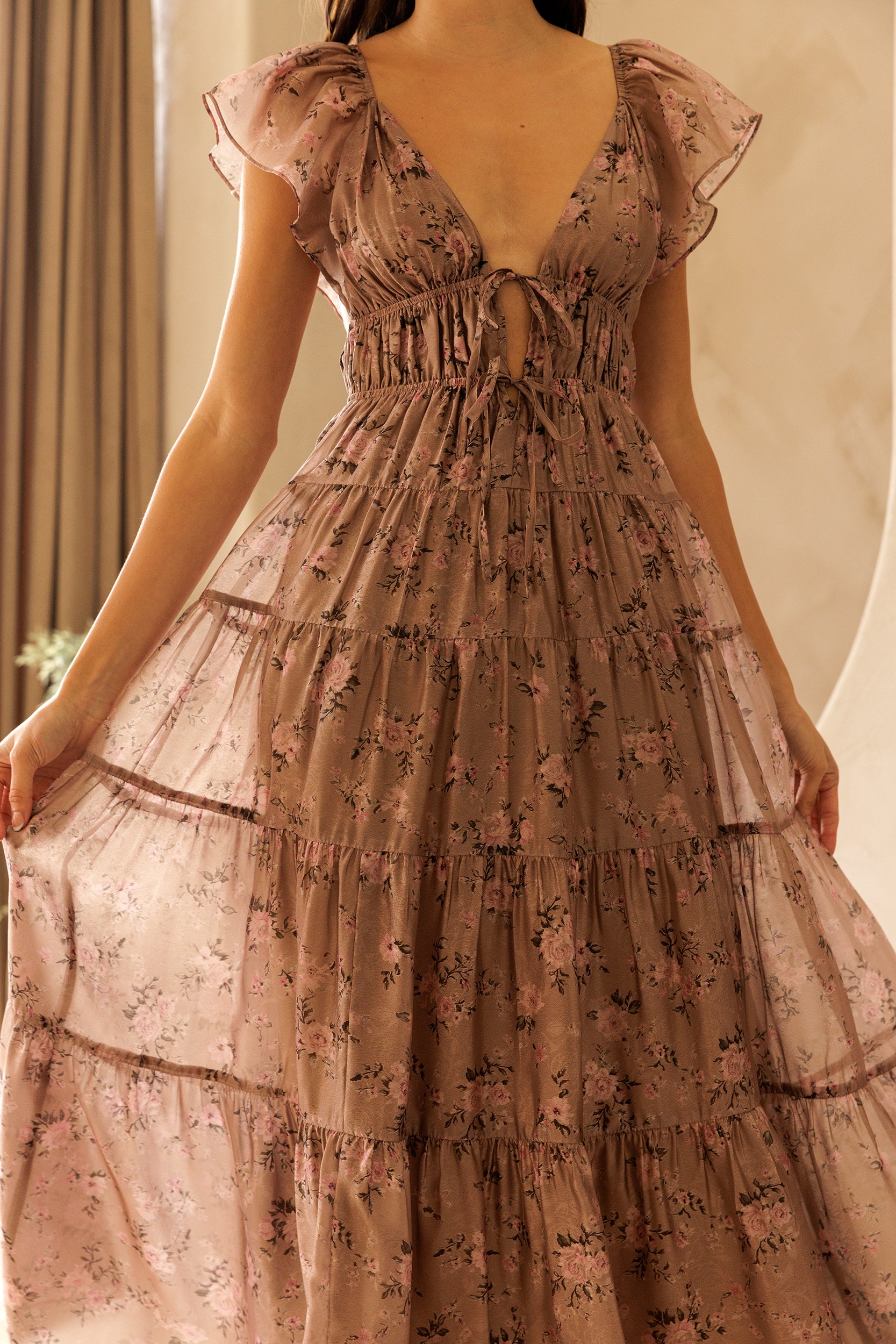 A person wearing a mauve midi dress with a floral pattern, flowy A-line silhouette, flutter sleeves and tiered skirt against a beige wall.