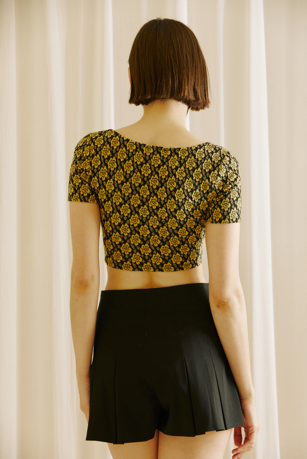 Back view of a woman wearing a black and yellow floral top and black shorts against a beige curtain background