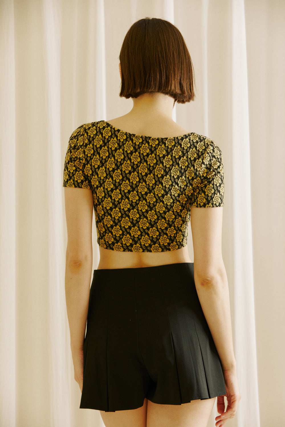 Back view of a woman wearing a black and yellow floral top and black shorts against a beige curtain background