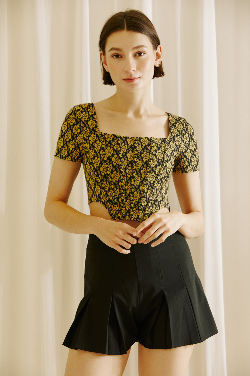 Woman wearing a floral top and black shorts against a beige curtain background