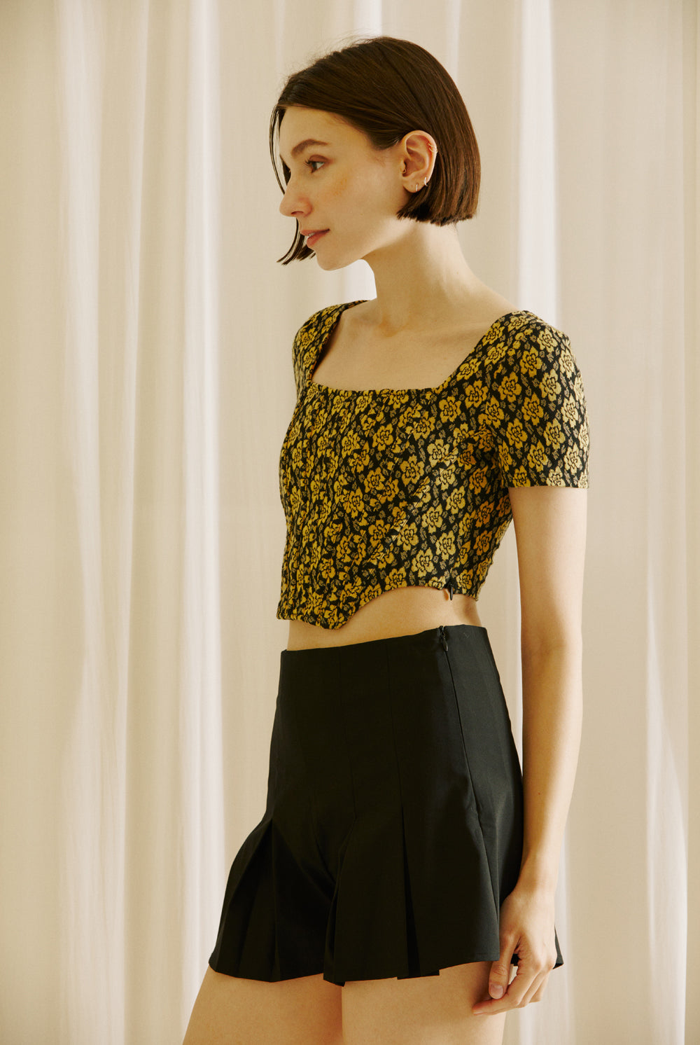 Side view of a woman wearing a floral top and black shorts against a beige curtain background