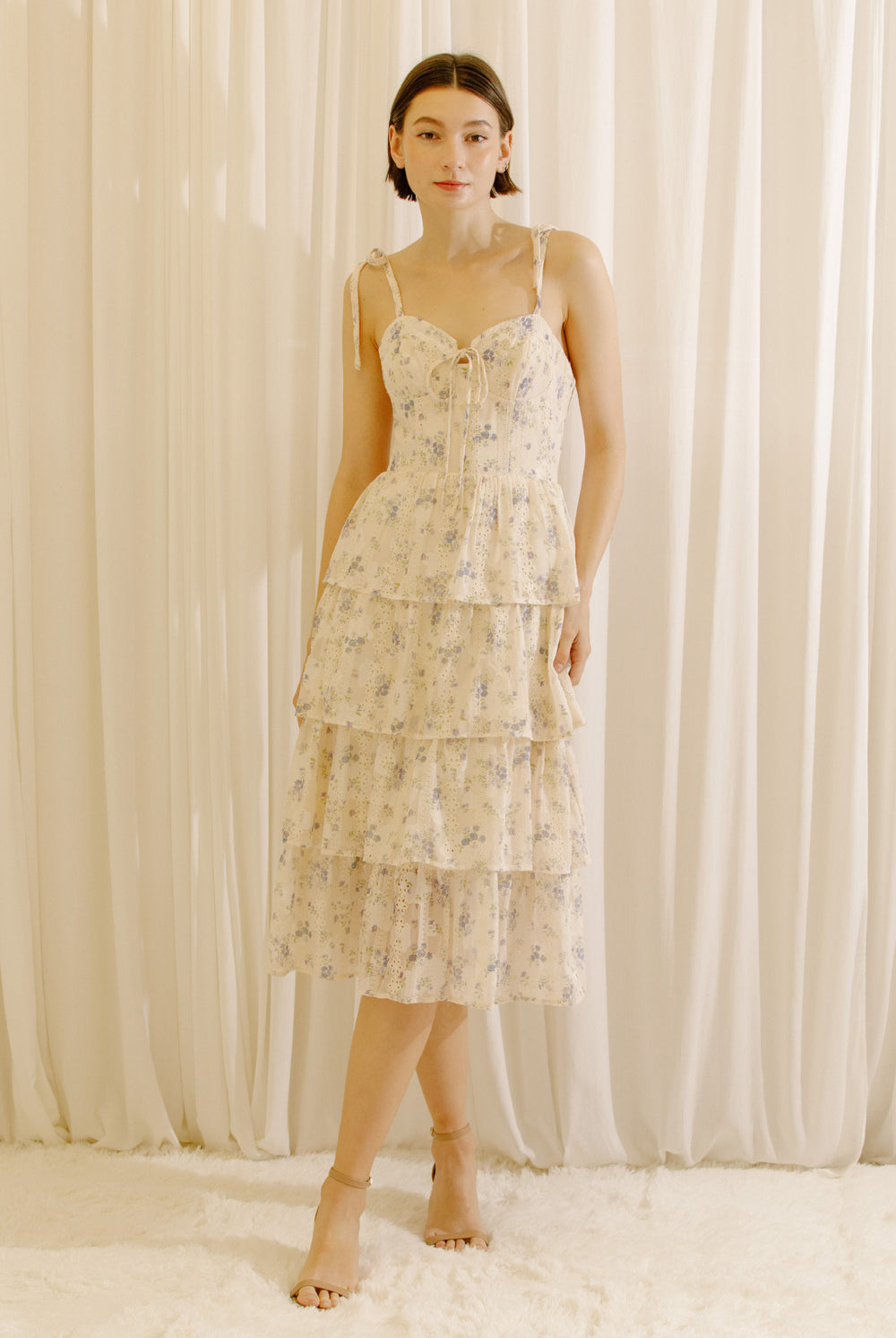 Woman wearing a cream floral midi dress with an eyelet design and a tiered skirt standing against a white curtain