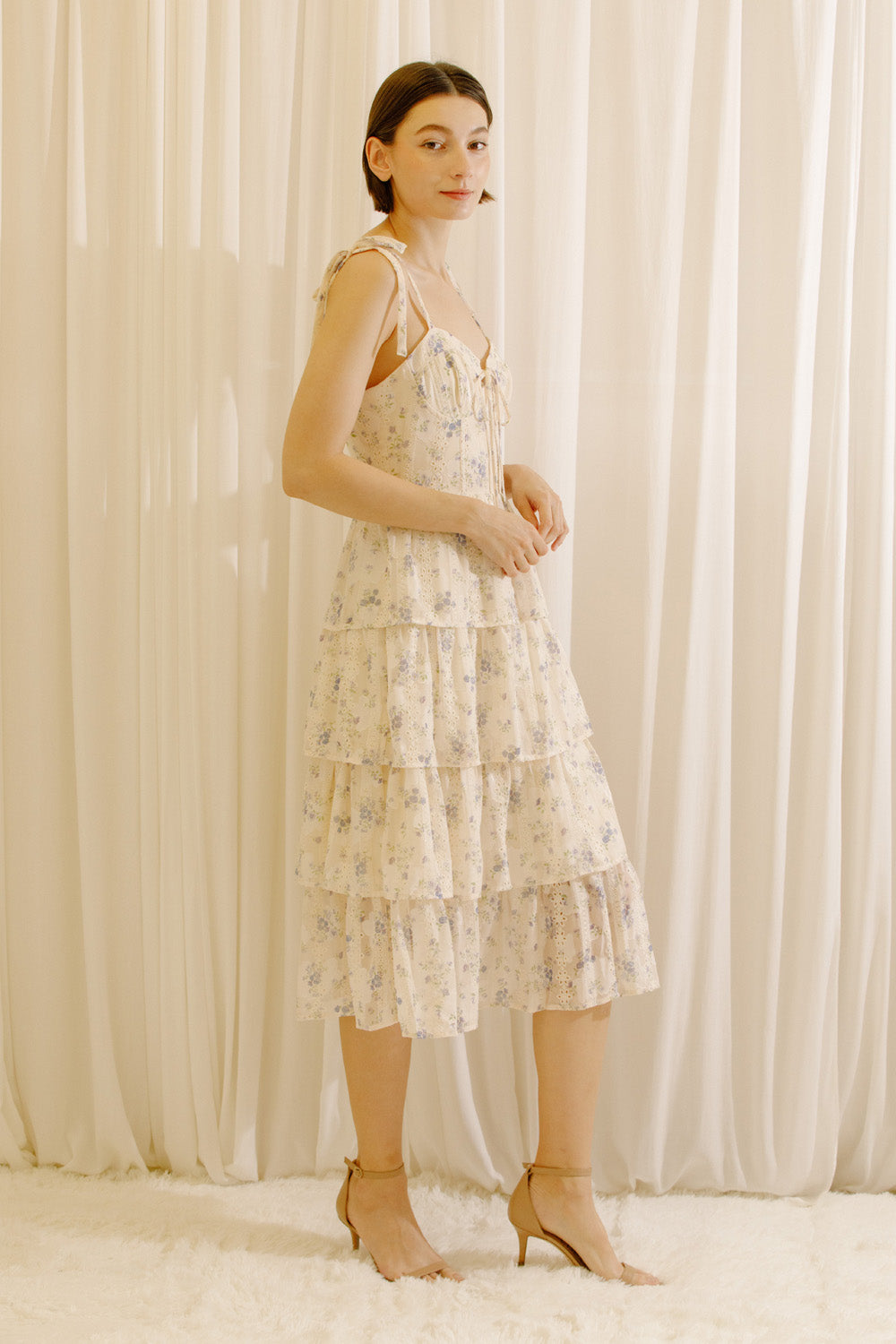 Side view of woman wearing a cream midi dress with a floral patter and eyelet design against a white curtain