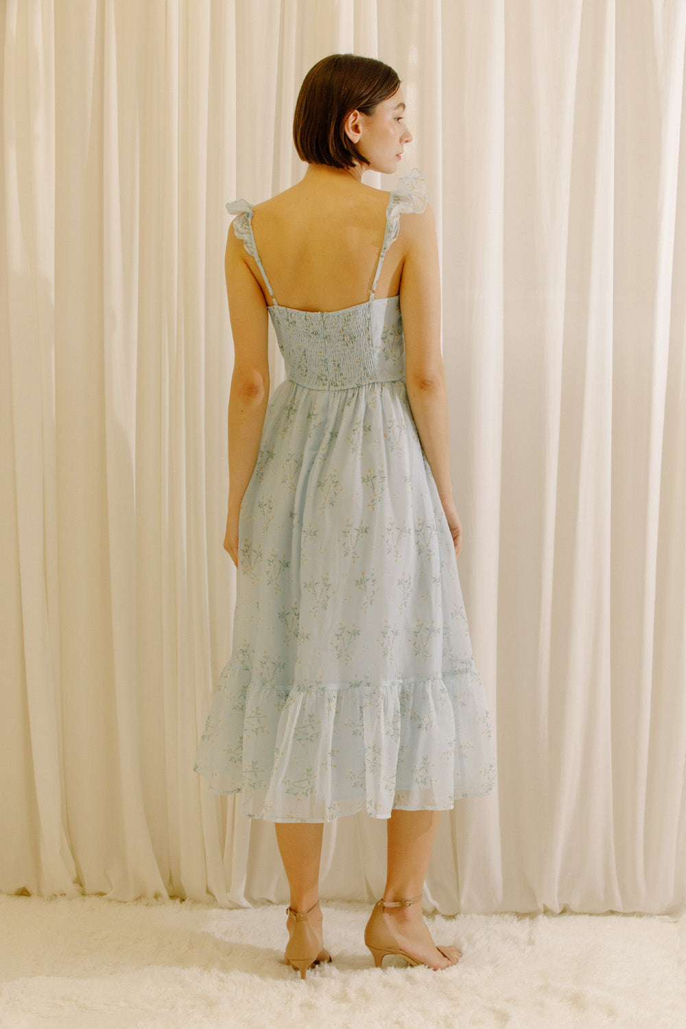 A woman stands with her back to the camera wearing a light blue tiered midi dress with ruffled shoulder straps and smocked upper back, set against a soft cream-colored curtain backdrop.