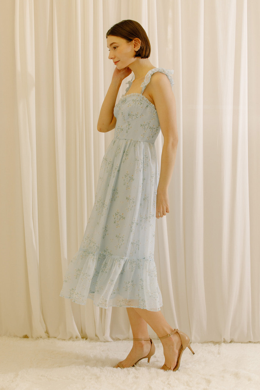 A woman wearing a light blue sleeveless midi dress with a delicate floral print, standing in front of a soft cream-colored curtain backdrop.
