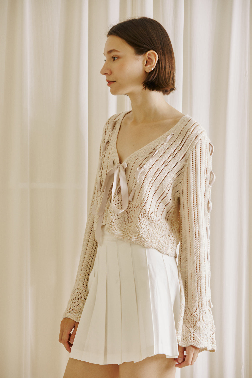 Side view of a woman wearing a beige knit cardigan with lace-up details and a white pleated skirt on a white background