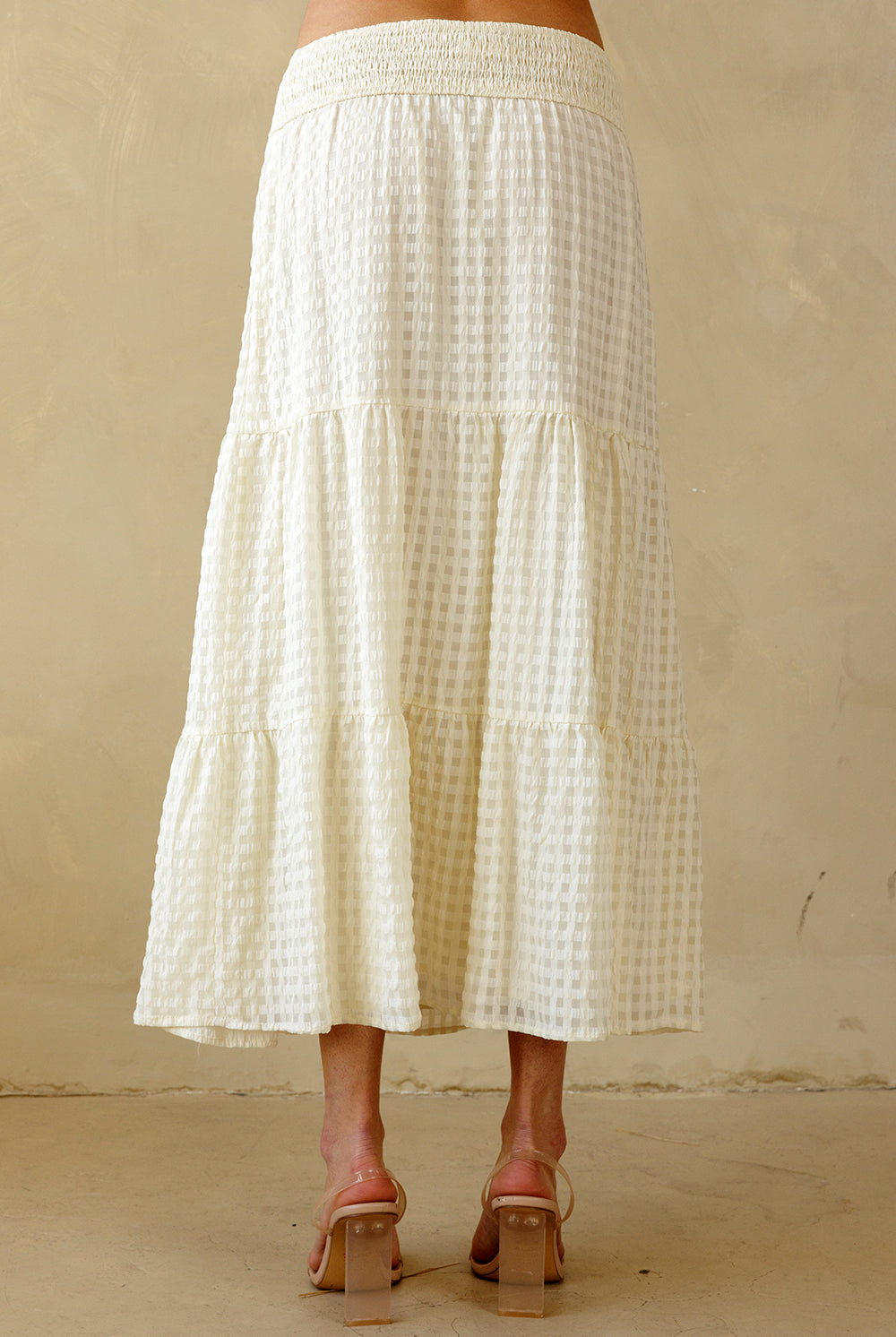 Back view of a cream maxi skirt with a subtle checkered pattern worn by a person on a beige background