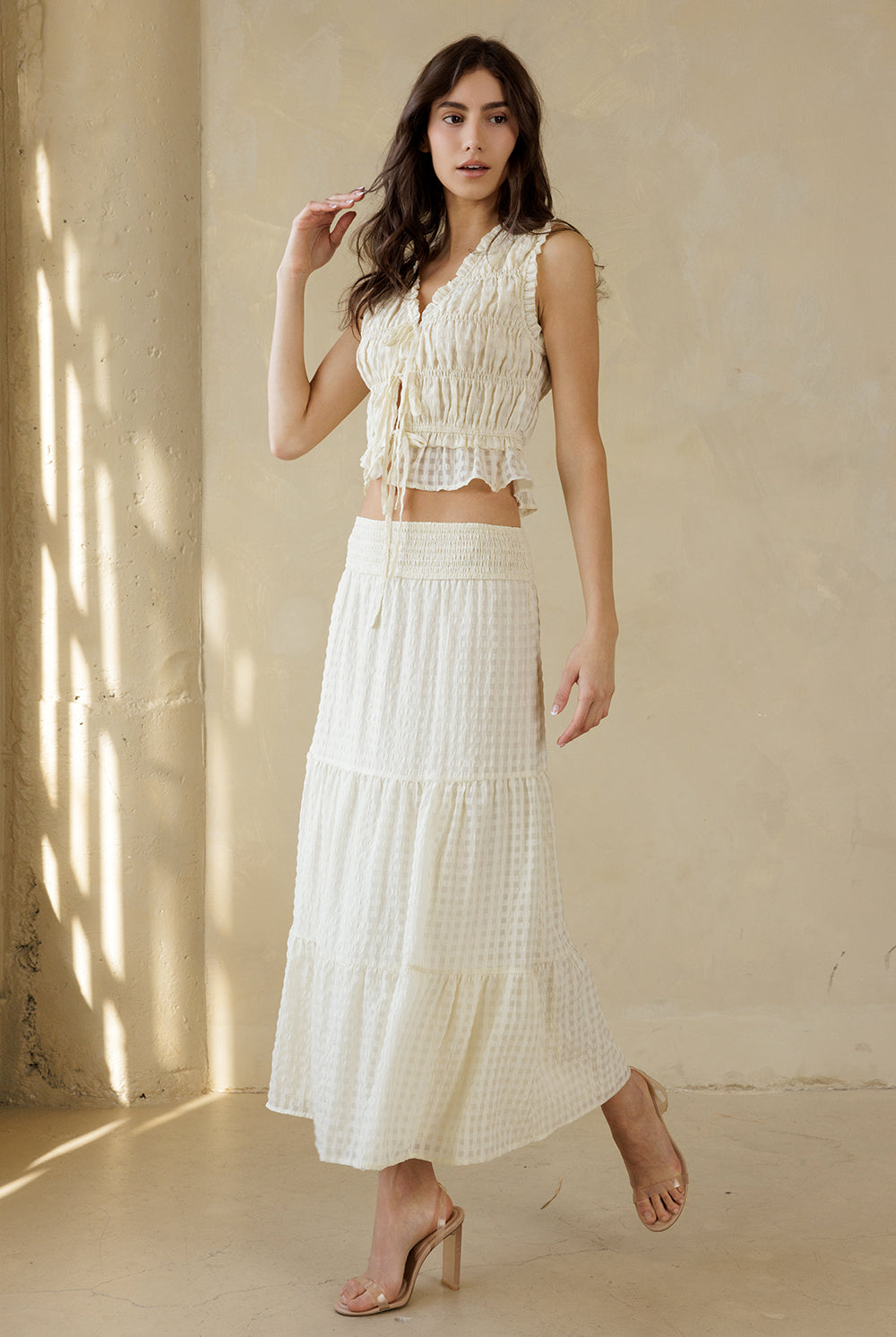 Woman wearing a cream textured maxi skirt with a subtle checkered pattern and a cream top standing against a beige wall with sunlight casting shadows.