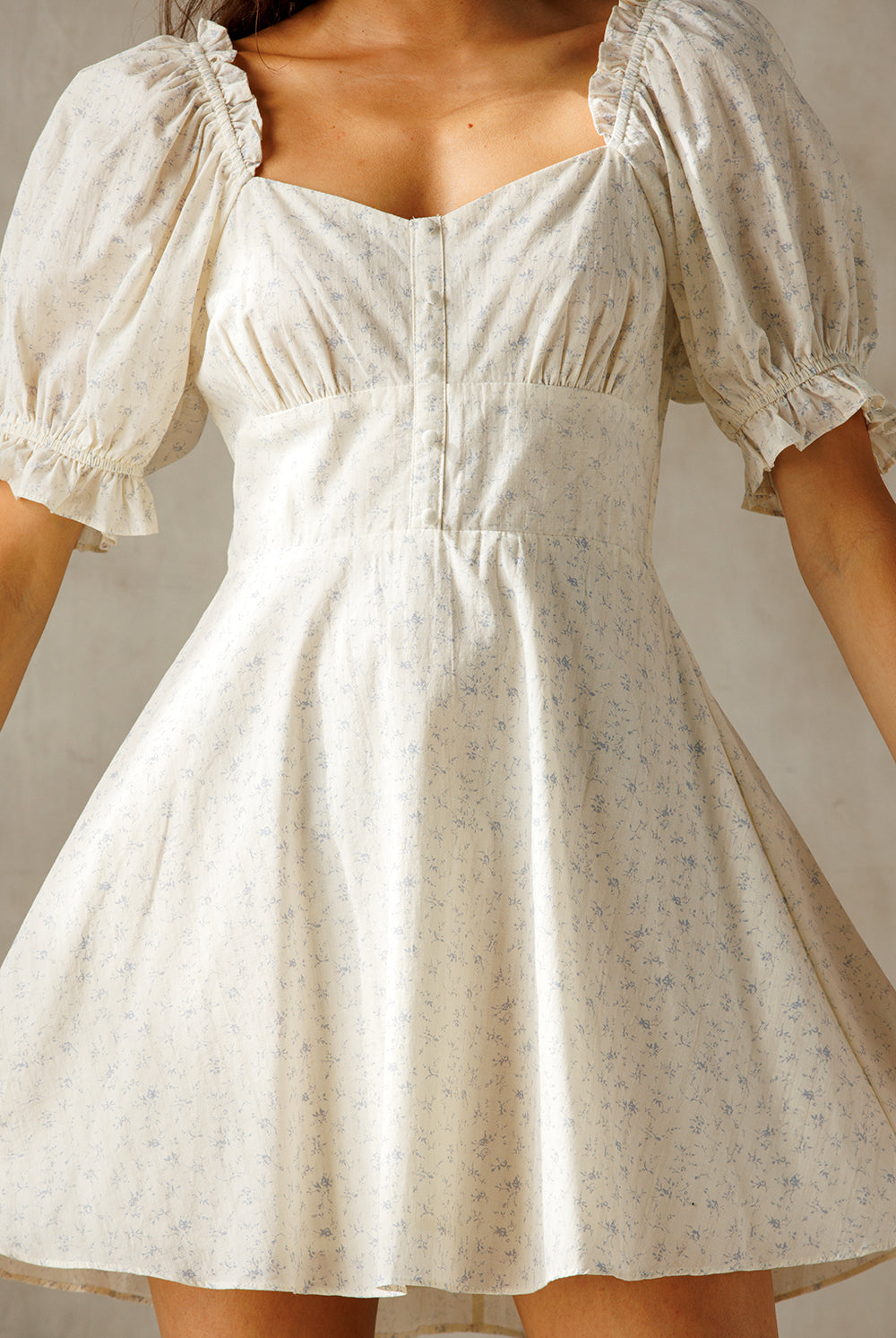 A woman wearing a short puff-sleeve dress with a square neckline and a flared skirt. The dress is cream with a subtle light blue floral print, photographed against a neutral light gray background.