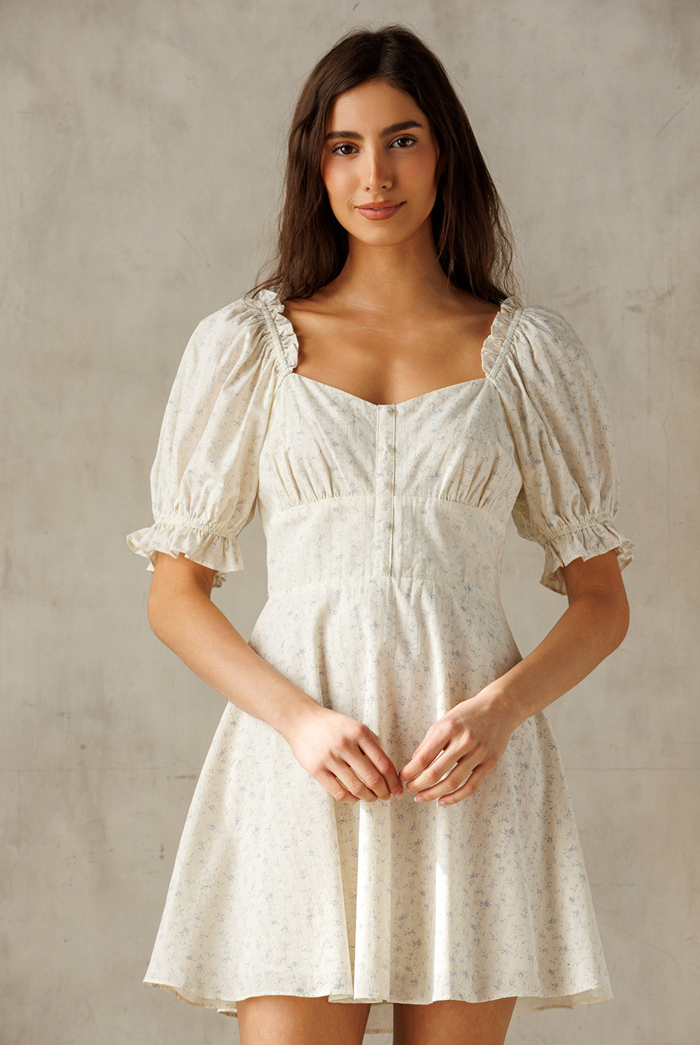 A woman modeling a short puff-sleeve dress with a square neckline, gathered bodice, and flared skirt in a cream fabric with a subtle floral print, set against a textured neutral background.