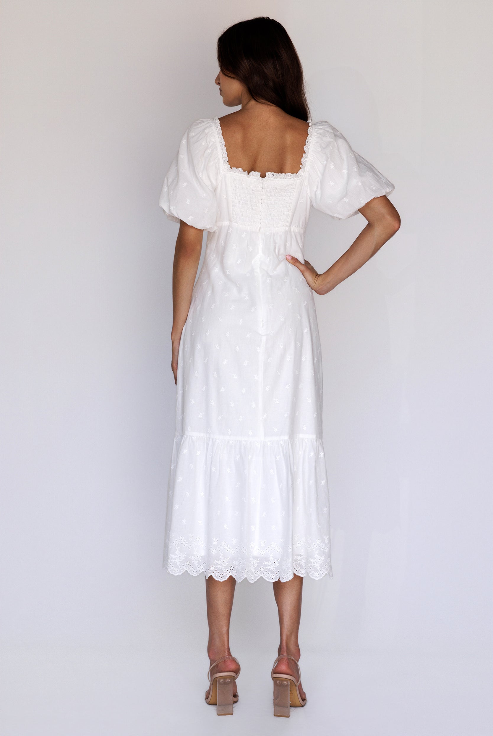A woman viewed from behind wearing a timeless white embroidered midi dress with puff sleeves, a smocked back, a back invisible zipper and scalloped eyelet hem.