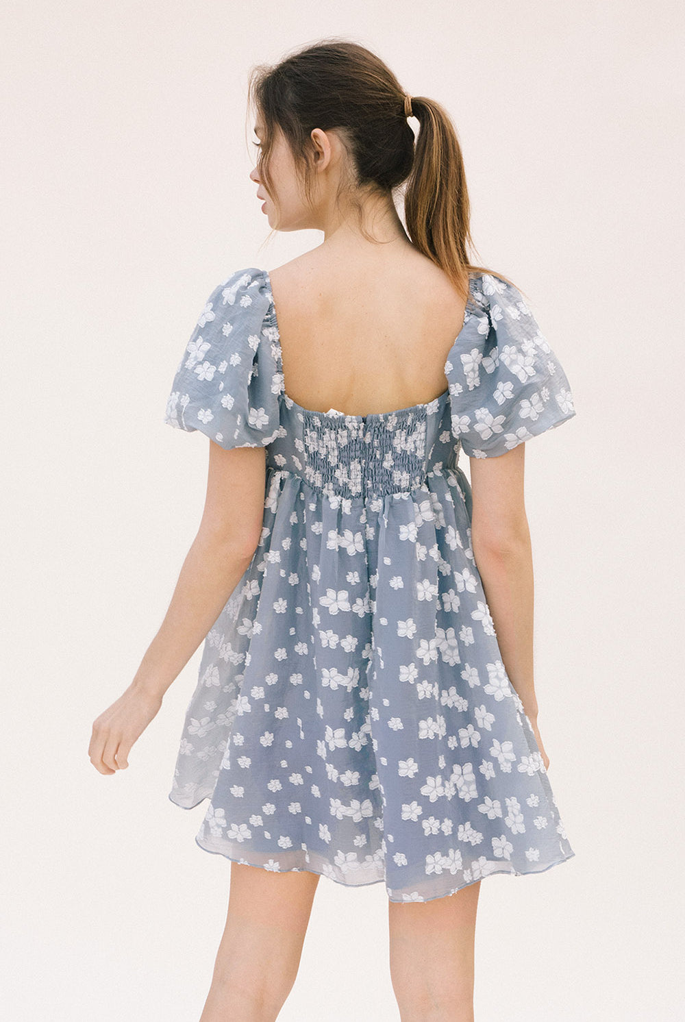 Back view of a woman wearing a blue dress with white floral embroidery, puff sleeves, back invisible zipper and smocked upper back on a white background