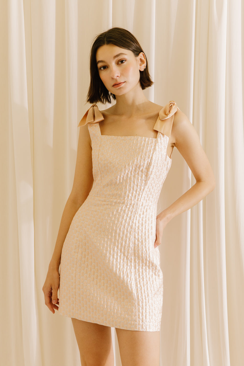 Woman wearing a pink dress with bow straps against a beige curtain background