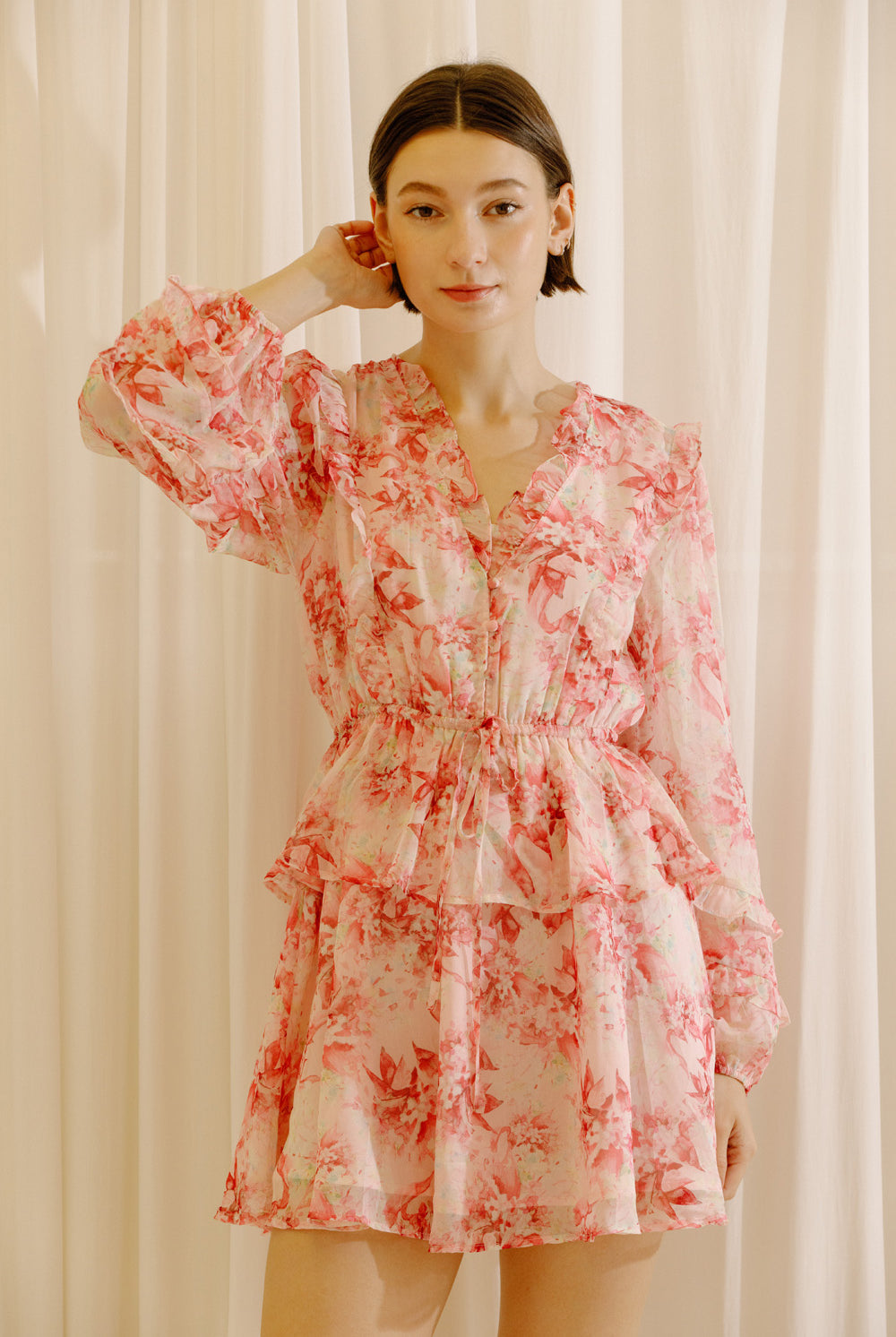 Woman wearing a pink and cream dress with floral print against a white curtain background