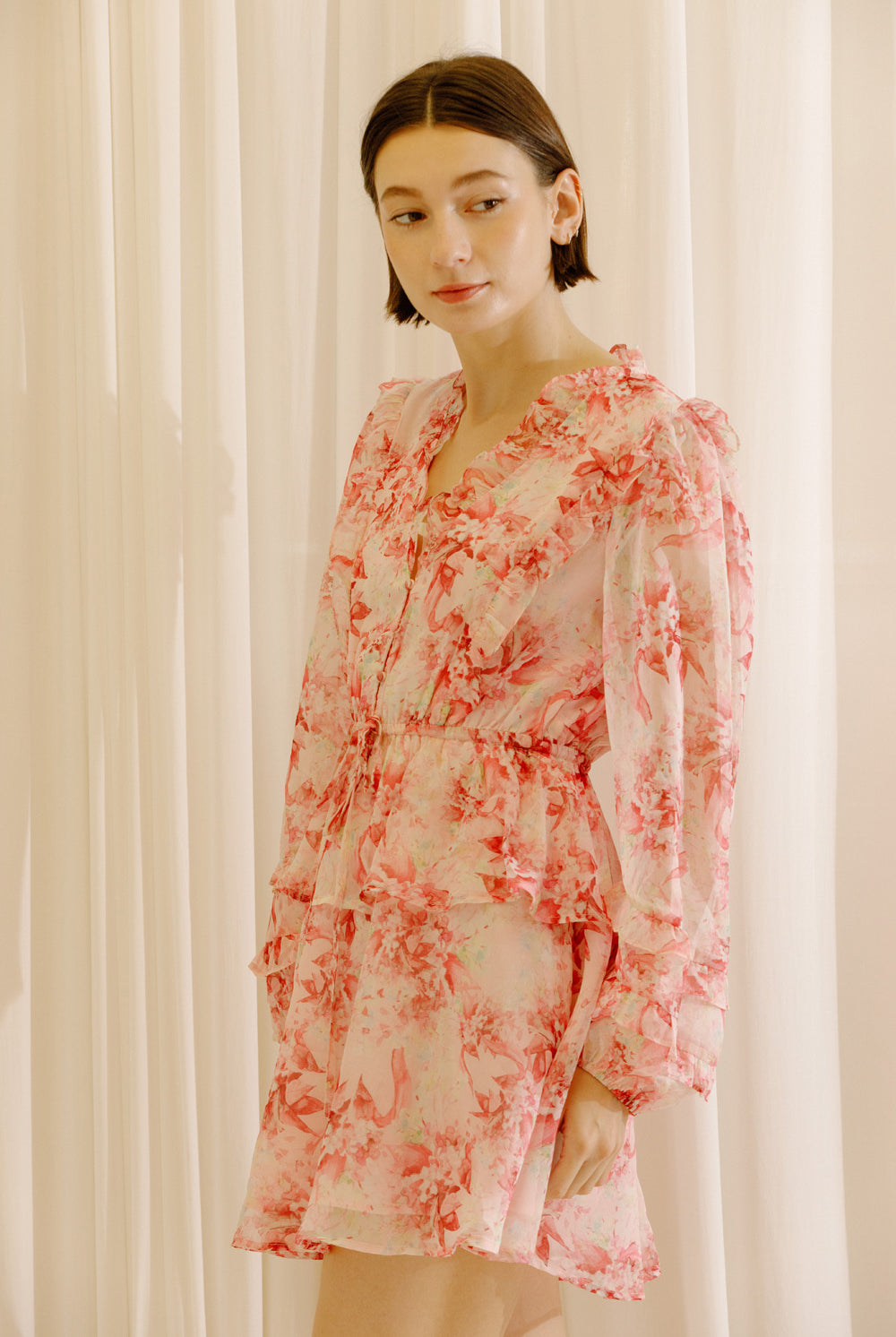 Side view of a woman wearing a pink and cream dress with floral print against a white curtain background
