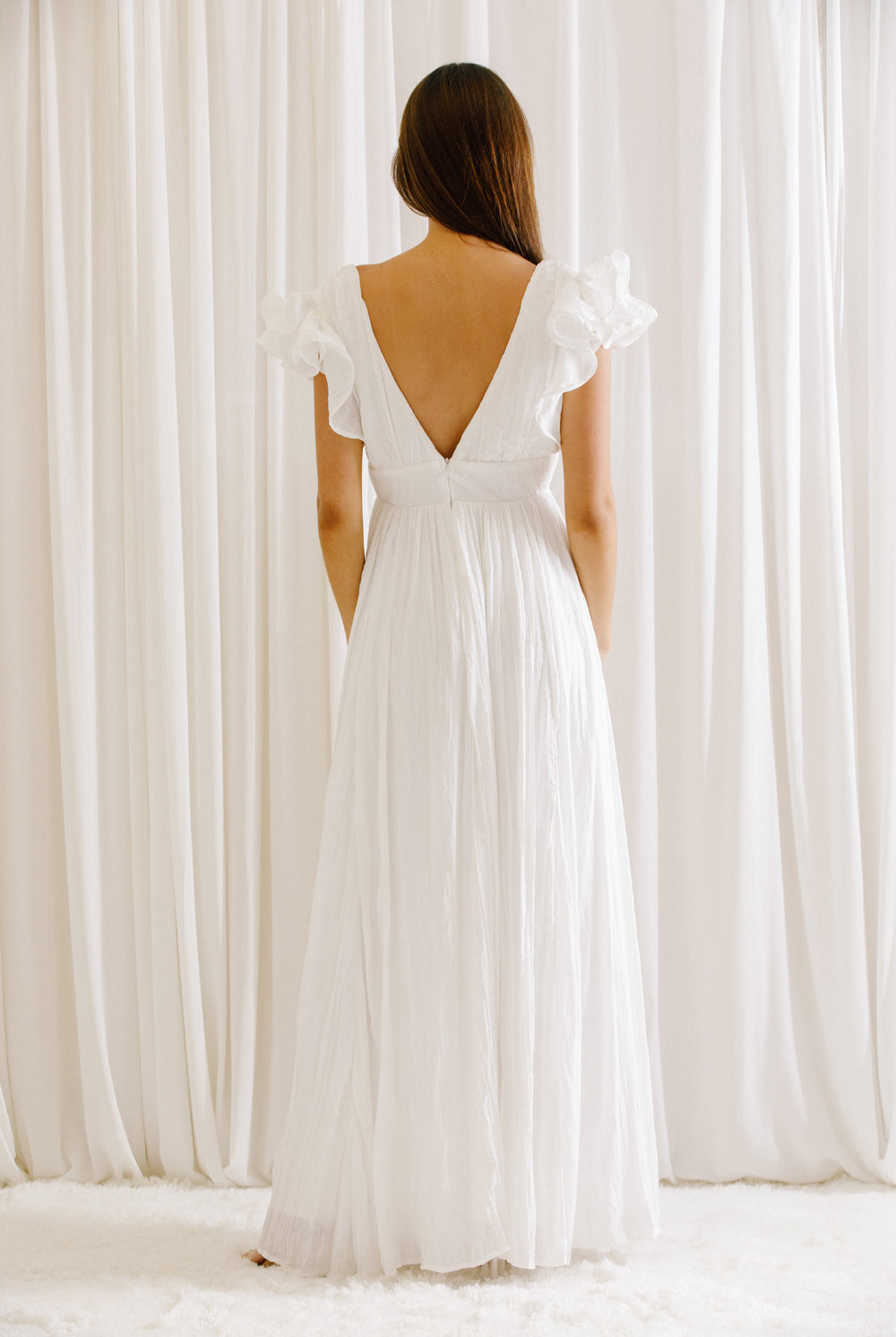 Back view of a woman wearing a white maxi dress standing against a white curtain